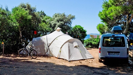 Een kampeerscène met een grote tent, een fiets en een geparkeerde auto omringd door bomen nabij een waterlichaam