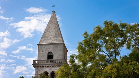A historic stone clock tower with a pointed roof, surrounded by green trees and a blue sky