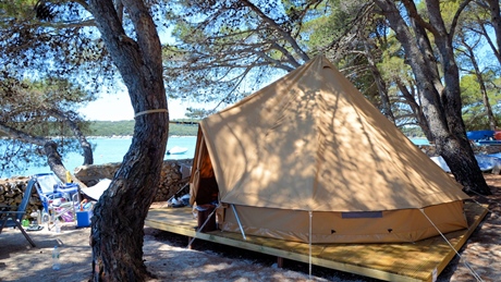 A large beige camping tent set up among trees with a view of the sea in the background