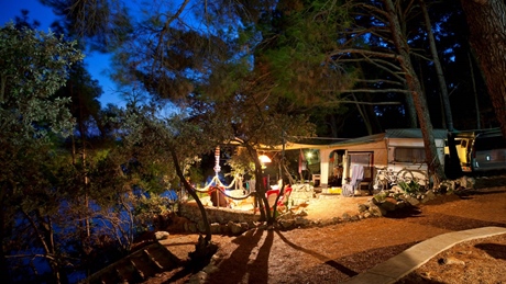 A scenic campsite set up in a forested area at night with a hammock, chairs, and a caravan