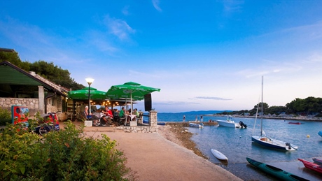 A seaside cafe with people dining under green umbrellas, overlooking small boats on the water