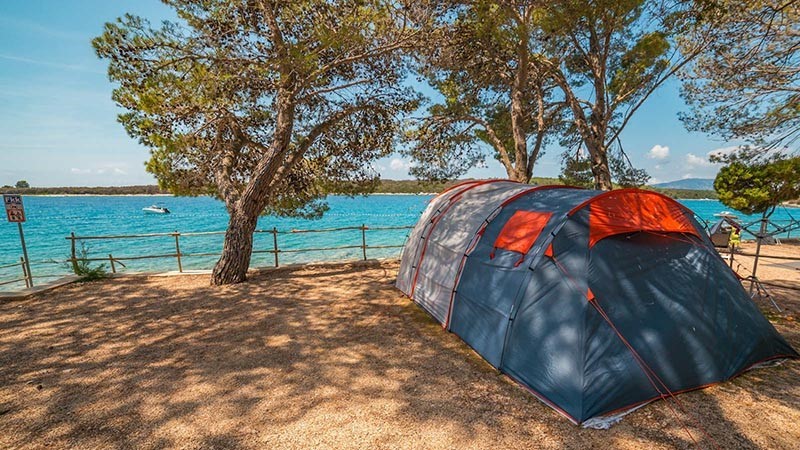 A camping tent set up near a scenic lakeside with trees and a clear blue sky