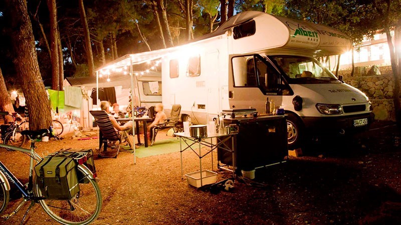 A camper van with the name Albergo Camper parked in a forest campsite, with people sitting at a table under a canopy