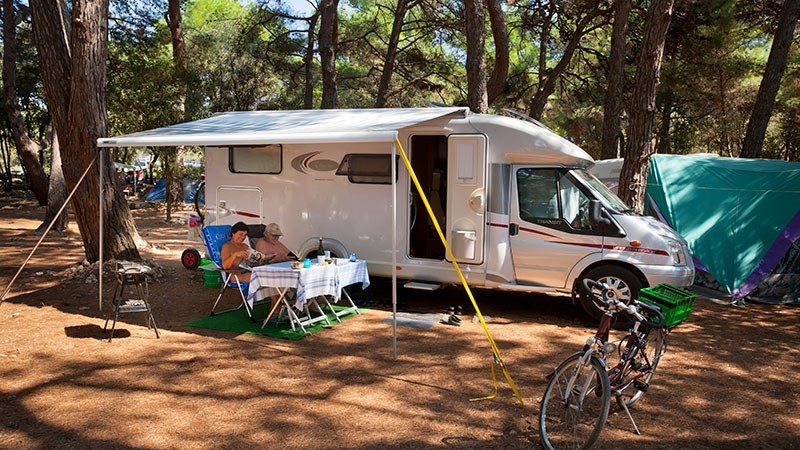 Una coppia si rilassa davanti a un camper in un campeggio boscoso, con una bicicletta parcheggiata nelle vicinanze
