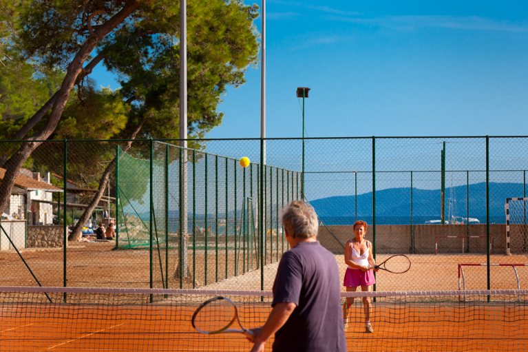 Two people playing tennis on an outdoor clay court near the beach
