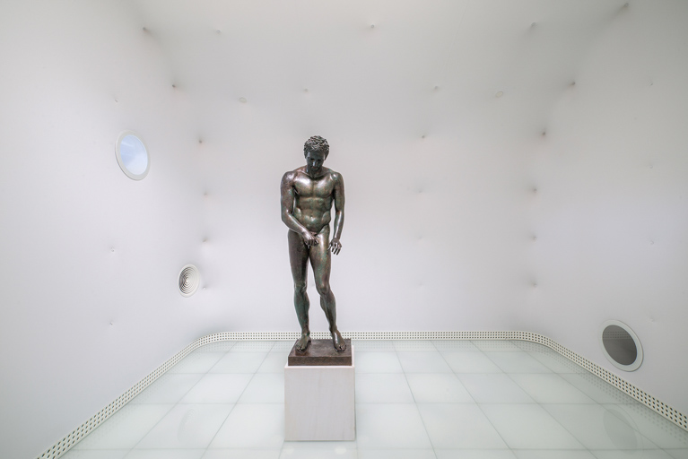 Bronze statue of a male figure displayed in a minimalist white room
